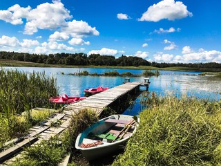 stara łódka nad brzegiem stawu pośród zielonych traw i niebieskiego nieba z białymi chmurami i starym mostem w tle