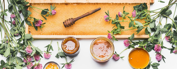 Otwarte słoiki miodu z czerpakiem, ramą o strukturze plastra miodu i dzikimi kwiatami na białym tle, widok z góry.