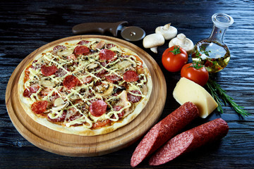 Gorąca pizza z salami, pieczarkami, pomidorami i serem na drewnianej desce. Gotowana pizza i surowi składniki na ciemnej drewnianej powierzchni.