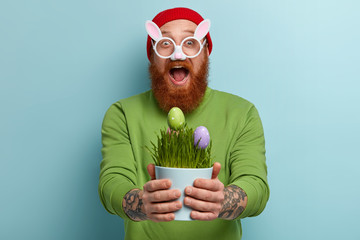 Joyful hipster wears bunny glasses, holds vase with paschal eggs as symbol of new life and resurrection, has fun coloring traitional food for Easter, meets spring holiday. Symbols and traditions