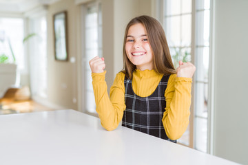 Beautiful young girl kid sitting on the table celebrating surprised and amazed for success with arms raised and open eyes. Winner concept.
