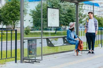 Couple communicating while waiting at bus stop. Hansome man and elegant woman with backpacks meets and talks at tram stop, outdoor. Students waiting for tram.