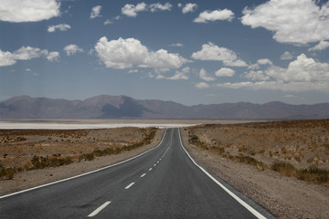 długa płaska droga asfaltowa ciągnąca sie po horyzont z andami i słonym jeziorem Salinas Grandes w tle