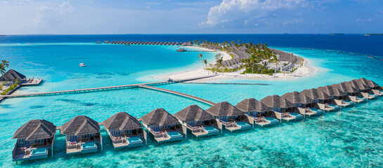 Aerial view of Maldives island, luxury water villas resort and wooden pier. Beautiful sky and ocean lagoon beach background. Summer vacation holiday and travel concept. Paradise aerial landscape pano