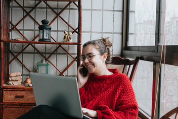 A young woman smiling while working on his laptop and making a call on a indie modern house