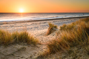 A sandy winding path weaves through the sand dunes and towards the sea on the Norfolk Coast at Winterton on Sea as the early morning sun rises above the horizon.
