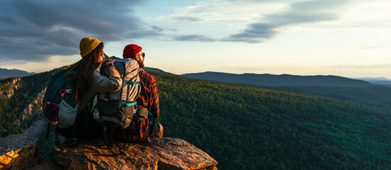 The couple meets the sunset in the mountains. Two travelers are sitting on the edge of a cliff and admiring the beautiful panoramic view. Travelers with backpacks at sunset in the mountains. Panorama