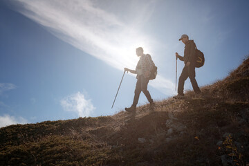Two men silhouettes walking along the top of the mountain with backpacks hiking gear meets the rising sun rays and blue sky background