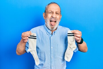 Handsome senior man with beard holding clean andy dirty socks sticking tongue out happy with funny expression.