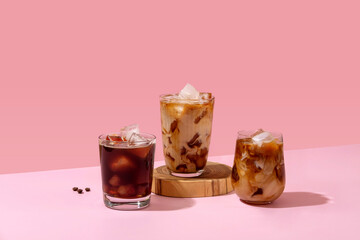 Ice coffee in a tall glass with cream poured over and coffee beans. Set with different types of coffee drinks on a pink table.