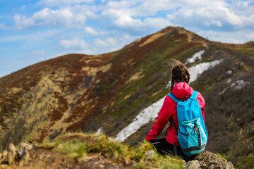beautiful hiker girl with backpack sitting on the grass with large colorful mountain in front of her; hiking in the mountains during early spring