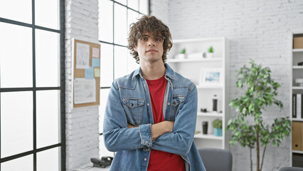 A young man with curly hair and a beard stands arms crossed in a bright modern office, exuding confidence.