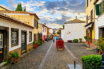 Tourists walk down the cobblestone Rua de Santa Maria narrow street of cafes, colorful doors and shops in the historic medieval old town of Funchal, Portugal, on the Canary Island of Madeira.