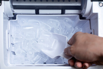 Closeup shot of a person's hand transferring bullet type ice cubes from a small portable ice maker into a clear glass with a small scoop.