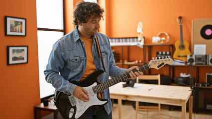 A young hispanic man plays guitar in a music studio with a modern interior design and musical equipment around him.