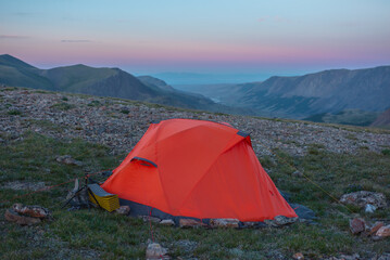 Orange tent on grassy stony pass with view to valley and mountain range silhouette far away under clear sunset color sky. Red tent close up on hill in high mountains under clouds in pure sunset sky.