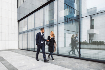A female leader walks in front of an office building in cloudy weather. She meets a male businessman. They are discussing a new deal. They are dressed in business clothes.