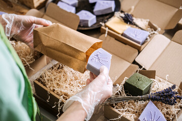 Person packs lavender soap into a kraft paper bag surrounded by shredded filler and handmade bars. Handmade product shipping, indie beauty dispatch, craft brand logistics, eco-packaging workflow