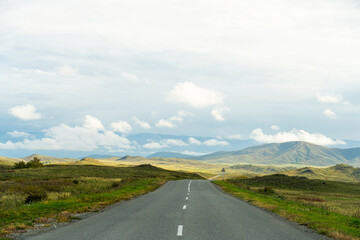 An empty paved road stretches into the distance, surrounded by expansive Altai mountain landscapes under a cloudy sky.