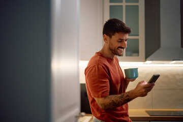 Happy man using mobile phone while drinking coffee in kitchen.