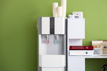 Modern water cooler with paper cups and workplace near green wall