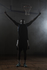 Basketball player standing on wooden court under hoop in dim gym while haze drifts around lights