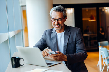 Smiling man using his smartphone while working on his laptop in an office