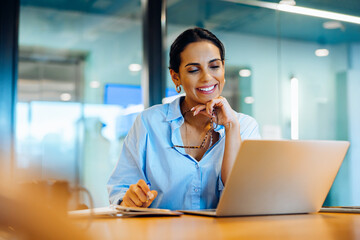 Smiling woman in office using laptop while holding glasses and working thoughtfully