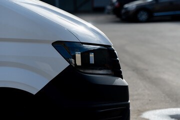 Close-up of modern white commercial van front headlight and black bumper, automotive design detail showing transport vehicle engineering, light reflection and functionality in urban environment