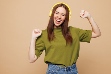 Young smiling happy woman wears olive green t-shirt casual clothes listen to music in headphones raise up hands dance isolated on plain pastel light beige background studio portrait. Lifestyle concept