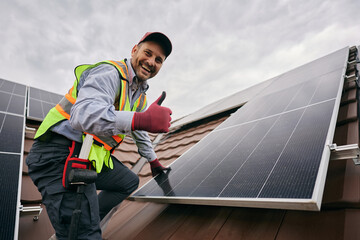 Happy worker showing thumb up while installing solar panels on roof and looking at camera