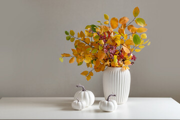 Bouquet of dry colorful red and orange fall leaves and branches in white vase and pumpkins on white table. Minimal modern composition. Autumn season. Home interior and season decoration.