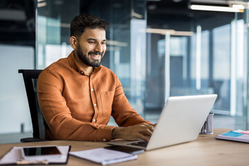 Young indian businessman smiling and typing on a laptop, sitting at a desk in a contemporary office reflecting productivity, success, and professional communication
