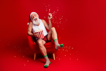 Santa enjoys popcorn on Christmas Eve relaxing in a red chair wearing hat striped socks and smiling