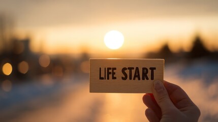 New Beginning: A person's hand holds a sign with LIFE START written on it. The sign is set against a serene backdrop of the setting sun, where the horizon meets the sky, creating a beautiful start.
