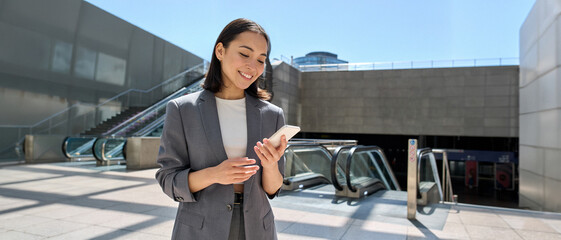 Young happy Asian business woman wearing suit holding mobile phone standing in city subway using smartphone for texting, checking apps for public transport, metro or travel guide