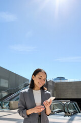 Young happy Asian business woman wearing suit holding mobile phone standing in city subway using smartphone for texting, checking apps for public transport, metro or travel guide