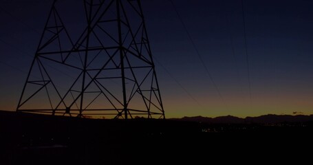 Dominating transmission tower looming left at dusk, sending power lines over ridge and town lights