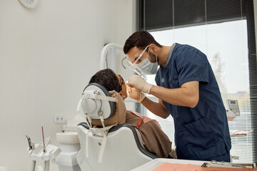 Young adult male dentist examining mature Caucasian woman sitting in dental chair during dental appointment, dentist wearing protective mask and gloves, dental equipment visible