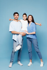 Full body young smiling fun happy parents mom dad with child kid girl wearing casual clothes hold daughter in hands look camera hugging isolated on plain blue background. Family day childhood concept.