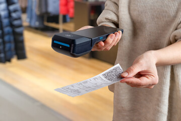 Hand offering printed receipt with QR code next to POS terminal inside clothing store, showing modern payment and checkout workflow