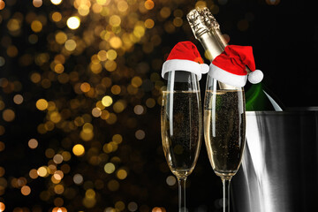 Sparkling wine in glasses with small Santa hats against blurred lights on black background