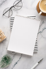 A stylish desk setup featuring a blank notebook on top of striped paper, glasses, a cup of coffee, a small wrapped gift, and seasonal decorations, perfect for holiday planning and creative work