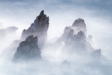 Long exposure landscape shot featuring jagged dark rock formations emerging from dense white mist or sea spray. High-contrast, minimalist aesthetic for editorial use.