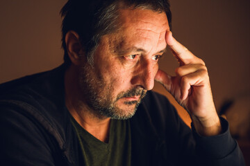 Concerned man looking at computer screen and thinking in dark room illuminated by warm low key light.