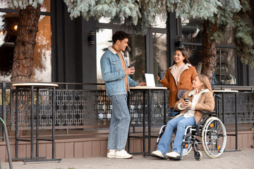 Young woman in wheelchair and her friends with coffee at street cafe