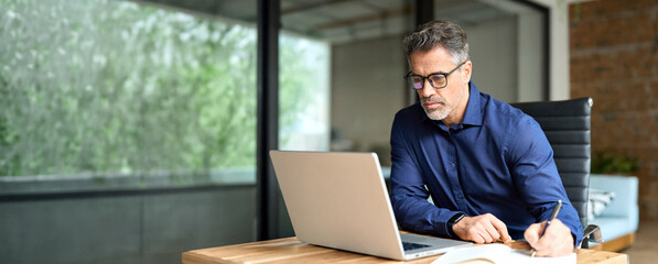 50 years old business man company manager investor working on computer technology sitting at office desk. Busy mature middle aged professional businessman executive using laptop at work writing notes.