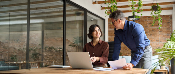 Happy busy executive leader working with colleague using laptop computer in office. Two professionals team people man and woman business partners talking on financial results at corporate meeting.
