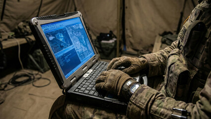 Military soldier using rugged laptop for tactical mission planning in field tent
