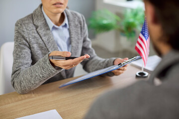 US visa interview at consulate, officer reviewing applicant documents at office desk with United States of America flag. Female lawyer with clipboard and man discuss USA immigration application